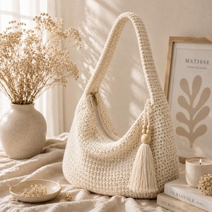 Può includere: Borsa a spalla all'uncinetto color crema con chiusura a zip e tracolla lunga. La borsa presenta un nappa con perline di legno. È appoggiata su un tavolo con un vaso di fiori secchi e una stampa incorniciata.