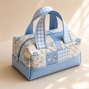 May include: A rectangular quilted fabric bag featuring a patchwork design of light blue floral patterns and blue and white gingham checks. The bag includes two matching gingham handles, a blue zipper closure, and a solid light blue polka dot base.