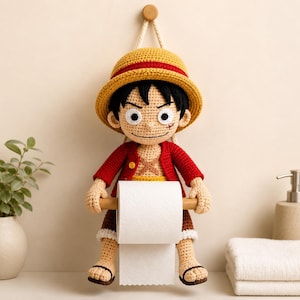 Op de afbeelding: Gehaakte Luffy toiletpapierhouder. Het personage draagt een gele strohoed met een rode band, een rode jas en een beige korte broek. De houder is bevestigd aan een houten deuvel die de toiletpapierrol vasthoudt. Het personage lacht.