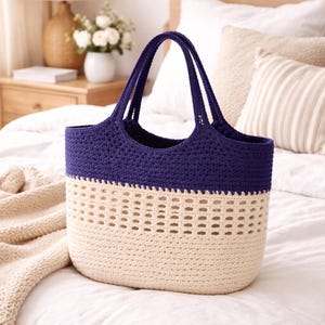 K&ouml;nnte beinhalten: Handgefertigte H&auml;keltasche in Creme und Marineblau. Oberteil und Henkel sind marineblau, der Korpus cremefarben mit einem dekorativen Lochmuster. Die Tasche liegt auf einer wei&szlig;en Bettdecke.