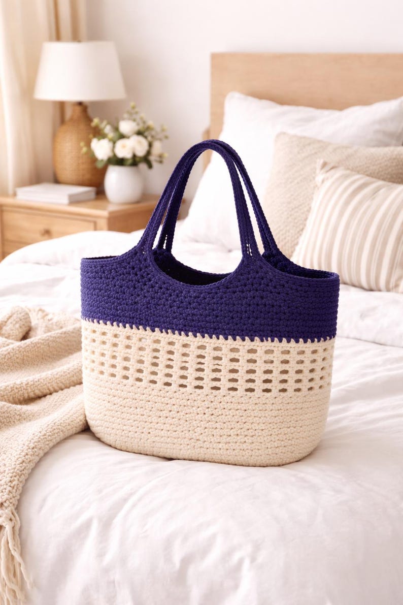 K&ouml;nnte beinhalten: Handgefertigte H&auml;keltasche in Creme und Marineblau. Oberteil und Henkel sind marineblau, der Korpus cremefarben mit einem dekorativen Lochmuster. Die Tasche liegt auf einer wei&szlig;en Bettdecke.