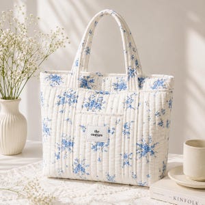 Puede incluir: Un bolso tote acolchado blanco con un estampado floral azul. El bolso tiene dos asas y un pequeño bolsillo en la parte delantera con el texto "the couture". El bolso está sobre una superficie blanca.