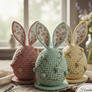 May include: Three pastel-coloured crochet Easter bunny decorations. The egg-shaped bunnies have large ears lined with floral fabric. Each bunny features embroidered floral details and sits on a round base, ideal for spring decorations.