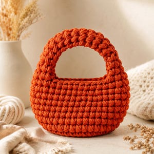 Puede incluir: Un pequeño bolso redondo de ganchillo naranja con un asa resistente. El bolso está hecho de hilo grueso, con una apariencia texturizada y tejida. El bolso está sobre una superficie neutra, con un jarrón y otros elementos decorativos en el fondo.