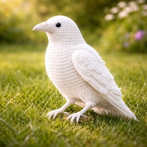May include: A handcrafted, white crochet bird with black eyes, perched on a small wooden branch. The bird's textured body and detailed wings are visible. The bird is set against a backdrop of green grass and soft sunlight.