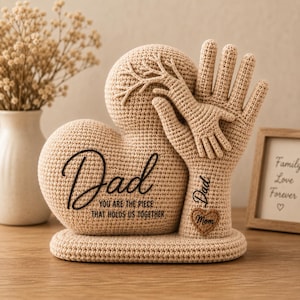 Könnte beinhalten: Eine beige Häkelskulptur in Herzform mit einer großen Hand, auf der eine kleinere Hand ruht. Der Text lautet Dad, You are the piece that holds us together, ergänzt durch ein kleines herzförmiges Patch mit der Aufschrift Mom und Dad.