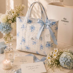 May include: A white quilted tote bag with a blue floral design, tied with a light blue ribbon. The bag has a tag that reads "Especially for you." The text "the couture" is on the bag. The bag is surrounded by flowers and cards. The bag has two handles.