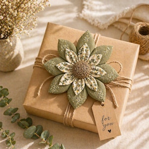 Puede incluir: Una caja de regalo envuelta en papel marr&oacute;n, adornada con una gran flor decorativa de tela en tonos verdes y crema. La caja est&aacute; atada con hilo de bramante, y una peque&ntilde;a etiqueta dice "for you" con un s&iacute;mbolo de coraz&oacute;n. Una ramita de eucalipto y un jarr&oacute;n con flores secas est&aacute;n en el fondo.