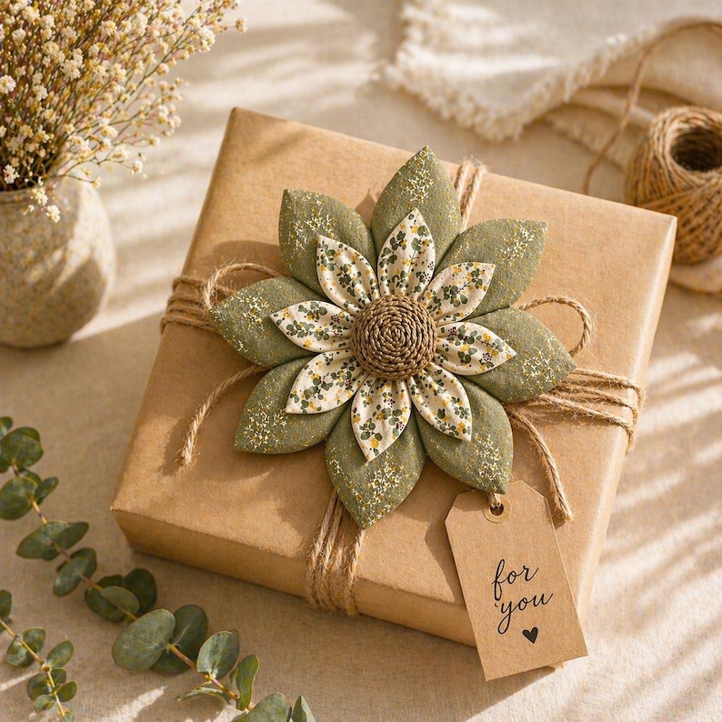Puede incluir: Una caja de regalo envuelta en papel marr&oacute;n, adornada con una gran flor decorativa de tela en tonos verdes y crema. La caja est&aacute; atada con hilo de bramante, y una peque&ntilde;a etiqueta dice "for you" con un s&iacute;mbolo de coraz&oacute;n. Una ramita de eucalipto y un jarr&oacute;n con flores secas est&aacute;n en el fondo.