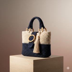 Può includere: Una borsa all'uncinetto bicolore blu navy e crema. La borsa presenta un manico blu navy, anelli di legno e una chiusura con fibbia in legno. Una nappa color crema pende dalla parte anteriore. La borsa è appoggiata su un cubo beige.
