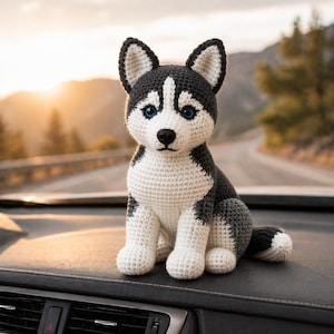 Może przedstawiać: Szydełkowa figurka psa husky z czarno-białym wzorem i niebieskimi oczami. Ręcznie robiona pluszowa zabawka siedzi na ciemnej powierzchni, z rozmytym tłem drogi i gór. Pies ma białą klatkę piersiową, łapy i pysk.