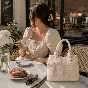 May include: A cream-colored woven handbag featuring two top handles and a decorative white tulle bow adorned with faux pearls. The bag has a structured rectangular shape with a vertical seam down the center and gold-tone hardware accents.