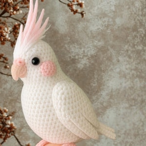 Può includere: Un uccello bianco all'uncinetto fatto a mano con cresta, guance e piedi rosa. L'uccello ha occhi neri e un becco color pesca. L'uccello artigianale è un oggetto decorativo, forse un giocattolo o un elemento di arredo per la casa.