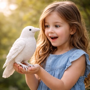 May include: A crocheted white bird with a long beak and black eyes. The bird is held in the hands of a young person wearing a blue denim dress. The background is a soft focus of green foliage and sunlight.