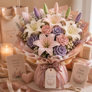 Könnte beinhalten: Ein handgefertigter Blumenstrauß aus gehäkelten Blumen in Rosa, Lila und Weiß. Der Strauß enthält Lilien, Rosen und andere florale Elemente, verpackt in rosa Papier und mit einem Band gebunden. Geschenk- und Grußkarten sowie ein kleines gehäkeltes Herz begleiten den Strauß.