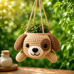 May include: A beige and brown crocheted hanging planter shaped like a puppy dog face. The planter features dark brown ears, a brown patch over one eye, a white muzzle, a black nose, and black eyes. It is suspended by three crocheted cords.
