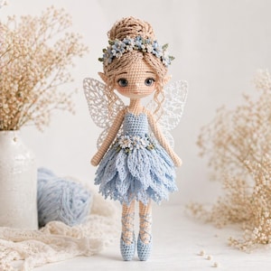 Op de afbeelding: Een gehaakte feeënpop met een lichtblauwe jurk en delicate witte vleugels. De pop heeft een bloemenkrans en balletschoentjes. Het haar van de pop is opgestoken in een knot. Een charmant, handgemaakt item.