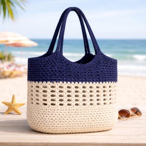 May include: A two-tone crochet tote bag with navy blue top and cream-colored bottom. The bag has two handles and a perforated design on the lower half. A starfish and sunglasses are on the wooden surface near the bag. The background shows a beach and ocean.