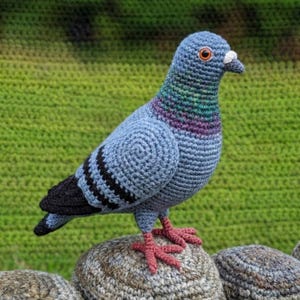 Puede incluir: Una paloma de ganchillo hecha a mano en tonos azules, grises y negros, posada sobre una piedra. El ave presenta costuras detalladas, ojos anaranjados y un pico blanco. Sus patas son de color rosa rojizo. El fondo es verde borroso.