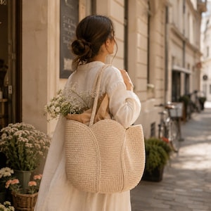 May include: A beige, woven tote bag with a tulip-shaped design. The bag is carried over the shoulder and is filled with flowers and a brown paper bag. The person is wearing a white dress.
