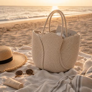 May include: A beige woven tote bag with two handles, sitting on a beach blanket. A straw hat with a black band, sunglasses, and sunscreen are also on the blanket. The background shows the ocean and a sunset.