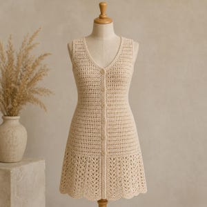 Può includere: Abito senza maniche all'uncinetto beige con scollo a V e abbottonatura frontale. L'abito presenta un corpetto aderente e una gonna leggermente svasata con un orlo traforato decorativo. L'abito è esposto su un manichino.
