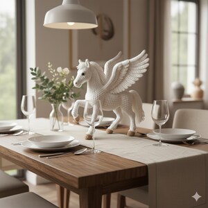 May include: A white crochet Pegasus figurine with outstretched wings, placed on a dining table set for a meal. The table is made of wood and has a beige runner, with white plates, glasses, and silverware. A vase of flowers is also on the table.