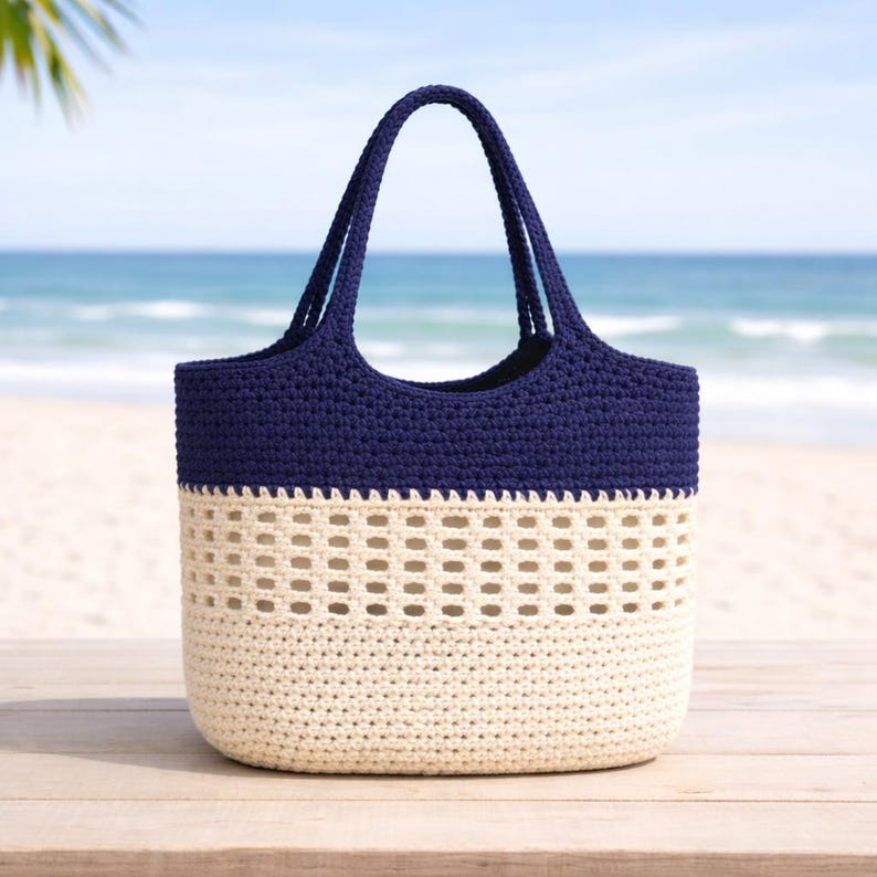 K&ouml;nnte beinhalten: Eine marineblaue und cremefarbene H&auml;keltasche mit zwei Henkeln. Der obere Teil und die Henkel sind marineblau, der untere Teil cremefarben mit einem Lochmuster. Die Tasche steht auf Holz, mit einem Strandhintergrund.