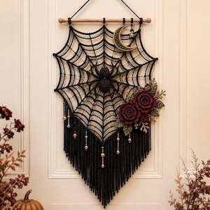May include: A black macrame wall hanging featuring a central spider on a web, accented with a gold crescent moon, a small star, and deep red crocheted roses with green leaves. The piece hangs from a wooden dowel with gold bead fringe accents.