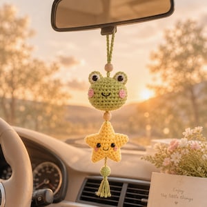 Puede incluir: Un adorno de coche hecho a mano, de ganchillo, con una rana verde y una estrella amarilla, ambas con caras sonrientes. La rana tiene mejillas rosas y la estrella una expresión linda. El adorno cuelga del espejo retrovisor de un coche.