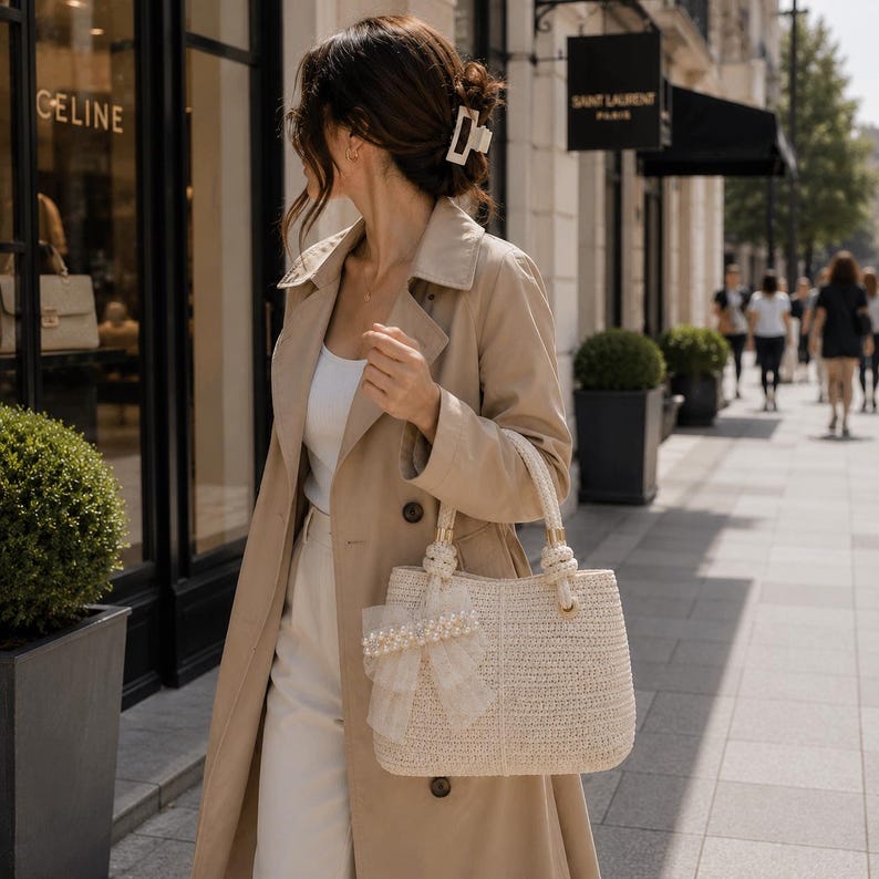 May include: A beige trench coat worn over a white top and white trousers, paired with a cream-colored woven handbag featuring a decorative tulle bow with faux pearls on the front and two braided handles, held by a person with a white hair clip.