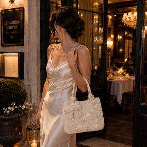 May include: A cream-colored satin slip dress with a cowl neckline and thin spaghetti straps, paired with a structured, woven straw tote bag featuring a decorative bow accent adorned with pearls and lace trim on the front panel.