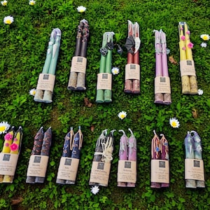 Op de afbeelding: Een assortiment decoratieve taps toelopende kaarsen in verschillende kleuren, waaronder groen, paars en geel, gerangschikt op een bed van groen gras met kleine witte bloemen. Elke kaars is gebundeld en gelabeld met een bruine papieren band.