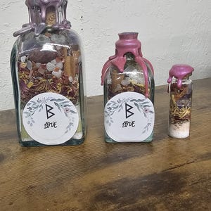 May include: Three glass bottles of varying sizes, filled with dried flowers, crystals, and other natural elements. Each bottle is sealed with a pink wax and has a label that reads "B Love". The bottles are arranged on a wooden surface.