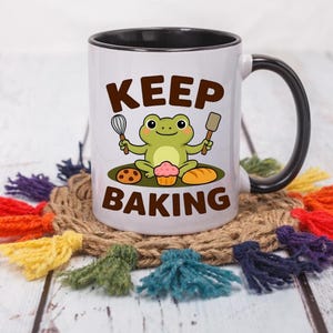 May include: White ceramic mug with a black interior and handle, featuring a cartoon frog holding a whisk and spatula, with the words "KEEP BAKING". The mug sits on a woven coaster with colorful tassels.