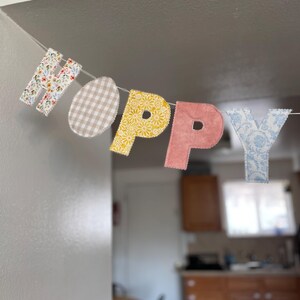Puede incluir: Una guirnalda decorativa con las letras "HOPPY" y una forma de huevo. Las letras y el huevo presentan estampados florales, cuadros vichy y damasco. La guirnalda est&aacute; ensartada en una fina cuerda blanca, perfecta para Pascua.