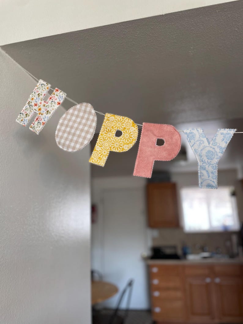 Puede incluir: Una guirnalda decorativa con las letras "HOPPY" y una forma de huevo. Las letras y el huevo presentan estampados florales, cuadros vichy y damasco. La guirnalda est&aacute; ensartada en una fina cuerda blanca, perfecta para Pascua.