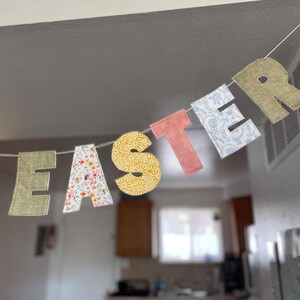 Puede incluir: Un cartel decorativo de Pascua que deletrea "EASTER" con letras de tela coloridas. Las letras presentan varios patrones, incluyendo dise&ntilde;os florales, amarillos y azules, ensartados en una cuerda blanca. El cartel est&aacute; colgado frente a un fondo borroso de cocina.