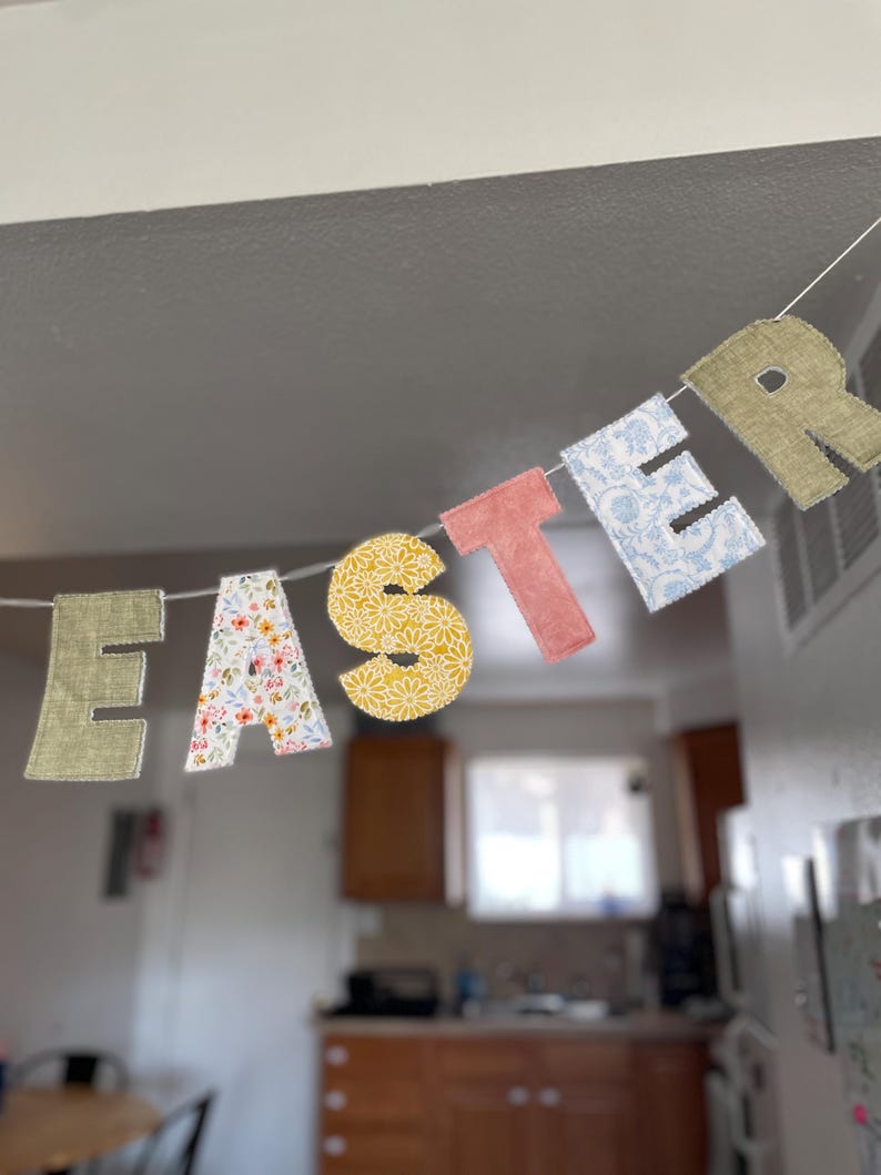Puede incluir: Un cartel decorativo de Pascua que deletrea "EASTER" con letras de tela coloridas. Las letras presentan varios patrones, incluyendo dise&ntilde;os florales, amarillos y azules, ensartados en una cuerda blanca. El cartel est&aacute; colgado frente a un fondo borroso de cocina.