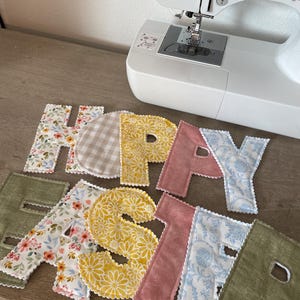 Puede incluir: Bandera hecha a mano "HAPPY EASTER", con letras de tela en dise&ntilde;os florales y estampados. Las letras est&aacute;n sobre una superficie de madera, cerca de una m&aacute;quina de coser. Las letras tienen bordes festoneados. Decoraciones de Pascua.