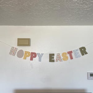 Puede incluir: Una guirnalda decorativa con la inscripci&oacute;n "HOPPY EASTER" en letras de tela coloridas y estampadas. La guirnalda est&aacute; colgada en una pared blanca, creando una decoraci&oacute;n festiva de Pascua. Las letras presentan dise&ntilde;os florales, cuadros y lisos.