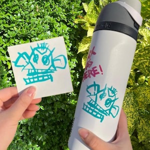 May include: A white water bottle with a teal blue graphic of a creature with a toothy grin. The text "Get It Here!" is written in pink on the bottle.