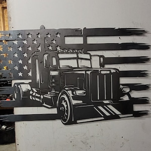 May include: A black metal cutout of a semi-truck in front of an American flag. The flag is distressed and has a faded look. The truck is detailed with a grill, headlights, and wheels.