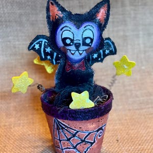 May include: A fabric bat figurine with a purple face, black wings, and a red bowtie, seated in a terracotta-colored pot painted with a black spiderweb design. The bat is surrounded by three yellow glittery stars attached to thin metal wires.