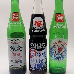 May include: Three green glass soda bottles with 7 Up and RC Cola logos. The bottles are souvenir bottles from the Indianapolis 500, the 1978 San Diego All-Star Game, and the State of Ohio. Each bottle has a different design and text.