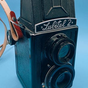 May include: A vintage black Lubitel 2 twin-lens reflex camera with a leather strap. The camera has two lenses and the name "Lubitel 2" is displayed on the front. The camera is set against a blue background.