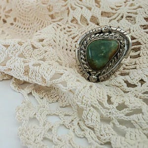Puede incluir: Un anillo plateado con una piedra verde ovalada grande. El anillo tiene un engaste detallado y texturizado y se muestra sobre un tapete de ganchillo color crema. La piedra tiene tonos marrones y verdes.