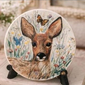May include: A decorative ceramic plate featuring a deer with large ears, surrounded by wildflowers and butterflies. The plate has a cream-colored background and is displayed on a black stand. The deer has brown fur and a black nose.
