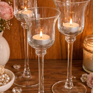 May include: Three clear glass candle holders with lit tea light candles inside. The candle holders have a tulip-shaped top and a long, slender stem. The background is a wooden surface with decorative elements.