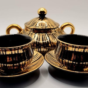 Pode incluir: Um conjunto de chá preto e dourado com duas chávenas com pires e um açucareiro com tampa. O conjunto tem um acabamento brilhante com riscas douradas verticais. As chávenas e o açucareiro têm pegas douradas.